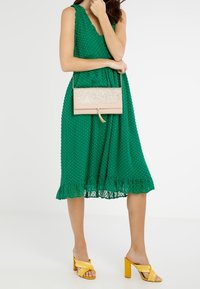 Vihreä pallokuvioinen mekko, jossa on röyhelöhelma, beige kimalteleva clutch-laukku ja keltaiset lohko- korkokengät ristikkäissuunnalla.