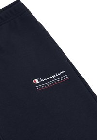 Dunkelblaue Jogginghose aus einem Baumwollmischgewebe. Mit einem weißen und roten Champion-Logo und dem Schriftzug "ATHLETICWEAR" am linken Oberschenkel.