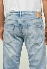 Vaqueros de mezclilla azul claro con una textura desgastada, que cuentan con bolsillos traseros con solapas angulares y una etiqueta de cuero en la cinturilla.