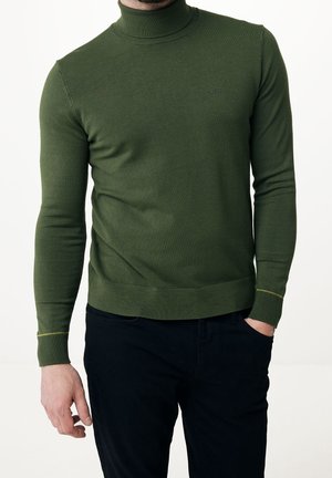 Homme portant un pull à col roulé vert olive avec un logo discret, pantalon noir, main gauche dans la poche, debout devant un fond blanc.