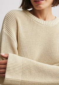 Femme portant un pull en maille épaisse crème avec col et poignets côtelés, tenant son bras opposé près du coude.