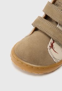 Világosbarna velúr cipő két állítható pánttal, gumitalppal, és díszítő varrással egy krémszínű panelen, piros árnyalattal.