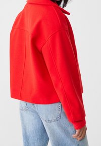 Veste en laine mélangée rouge avec un col et des épaules tombantes. Présente des poignets côtelés et une coupe carrée. Associée à un jean bleu clair.