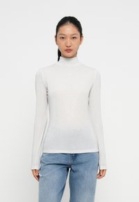 Jauna sieviete ar melniem matiem sasietiem atpakaļ, ģērbusies baltā garajā piedurknē turtleneck krekli un gaiši zilās nolietotajās džinsos, stāv uz vienkrāsaina baltā fona.