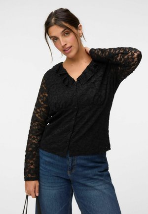 Frau trägt eine schwarze Spitzenbluse mit langen Ärmeln und Knöpfen sowie blaue Jeans, hält eine schwarze Umhängetasche und posiert mit einer Hand am Hals.