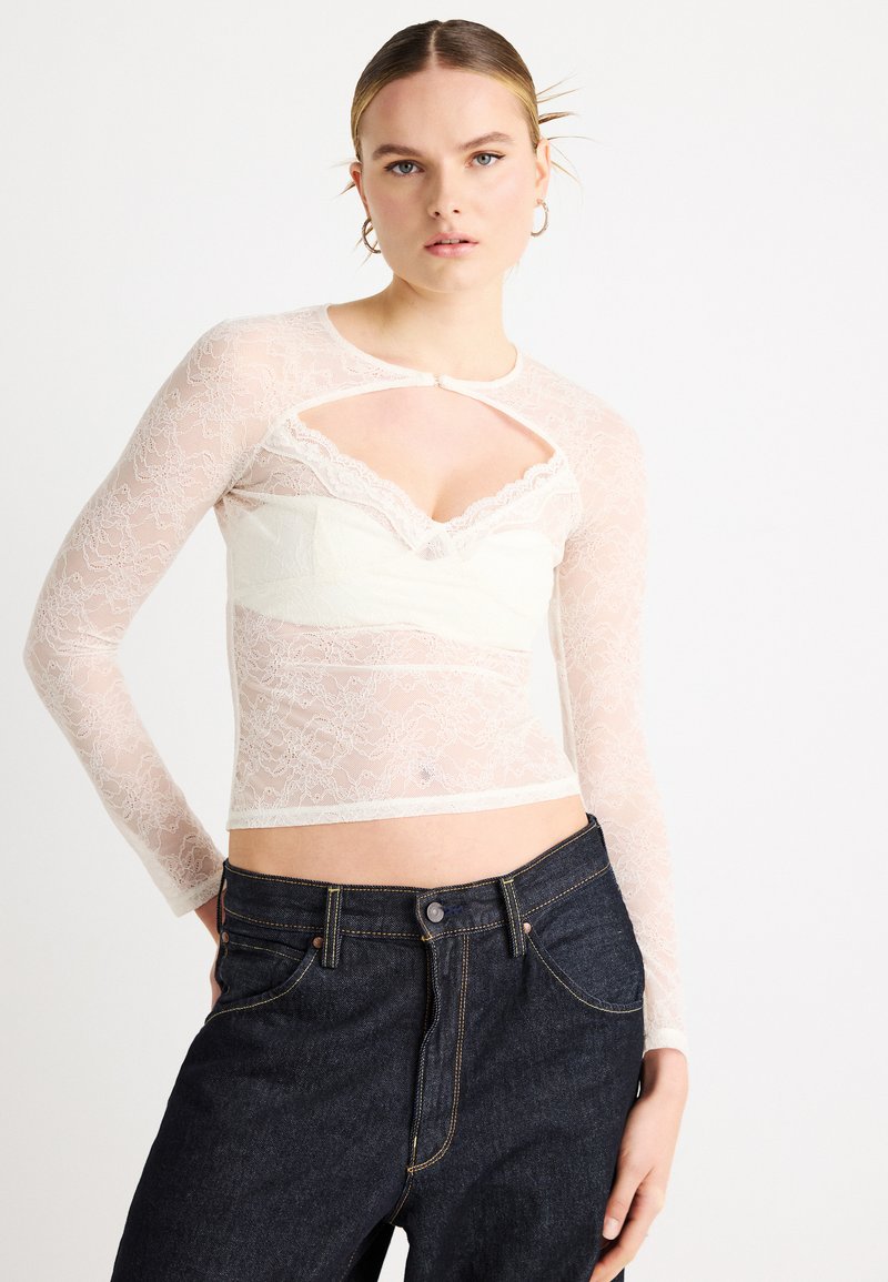 Λευκό μακρυμάνικο κορμάκι από δαντέλα με άνοιγμα τύπου keyhole και περιθώριο με κυματιστό σχέδιο, φορεμένο πάνω από λευκή υφασμάτινη βάση. Συνδυασμένο με σκούρο τζιν.