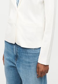 Personne portant un blazer blanc sur une chemise à rayures bleu et blanc, associée à un jean en denim bleu clair, debout les mains le long du corps.