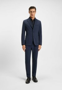 Marine blauwe pak met een getailleerde pasvorm, voorzien van een eenknoopssluiting jasje, twee voorkooien en bijpassende broeken. Zwarte nette schoenen maken de look compleet.