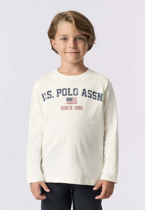 Camicia a maniche lunghe in cotone color crema con testo "U.S. POLO ASSN." e grafica di una bandiera in blu navy e rosso, indossata con pantaloni scuri.