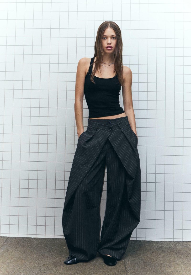 Zwarte tanktop gecombineerd met brede, donkergrijze pantalon met streepjespatroon, featuring een unieke gedrapeerde design en twee zijzakken.