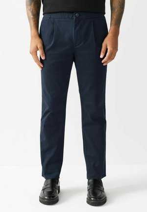 Navy blue trousers with a relaxed fit, front pleats, elastic waistband, and a button closure, paired with black slip-on shoes.