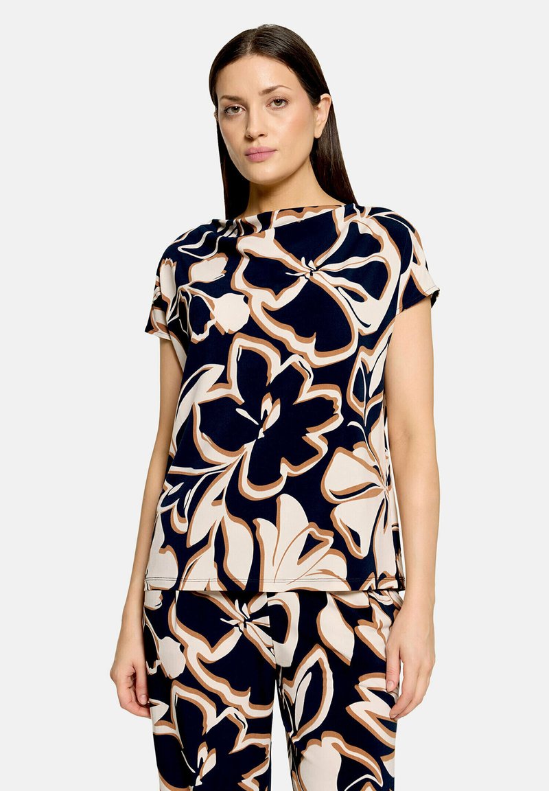 Femme aux cheveux lisses et foncés portant un haut et un pantalon assortis avec de grands motifs floraux abstraits bleu marine, crème et beige, debout devant un fond uni.