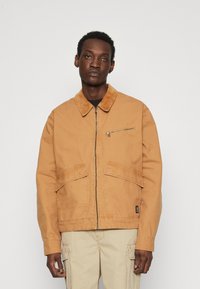 Tan bomuldjakke med en kordfløjelkrave, frontlynlås og to store frontlommer. Designet har en ensfarvet overflade med minimal detaljering.
