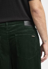 Groene corduroy broek met een achterpatch met logo, textuurribbels en twee achterzakken. Zwarte shirt gedeeltelijk zichtbaar boven.