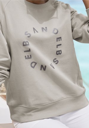 Beige Sweatshirt mit dem Aufdruck "SANDELBELD" in einem kreisförmigen Muster auf der Vorderseite, kombiniert mit weißen Hosen vor einem hellblauen Hintergrund.