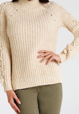 Pull beige en maille avec un col montant et des manches décoratives à lacets, porté avec un pantalon vert olive.