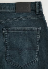 Mörkblå denimjeans med en strukturerad yta. Har en läderpatch på midjan och en enda bakficka med kontrasterande sömmar.