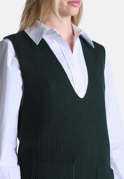 Femme portant un gilet sans manches en maille vert foncé par-dessus une chemise blanche à col boutonné, vue des épaules aux hanches.
