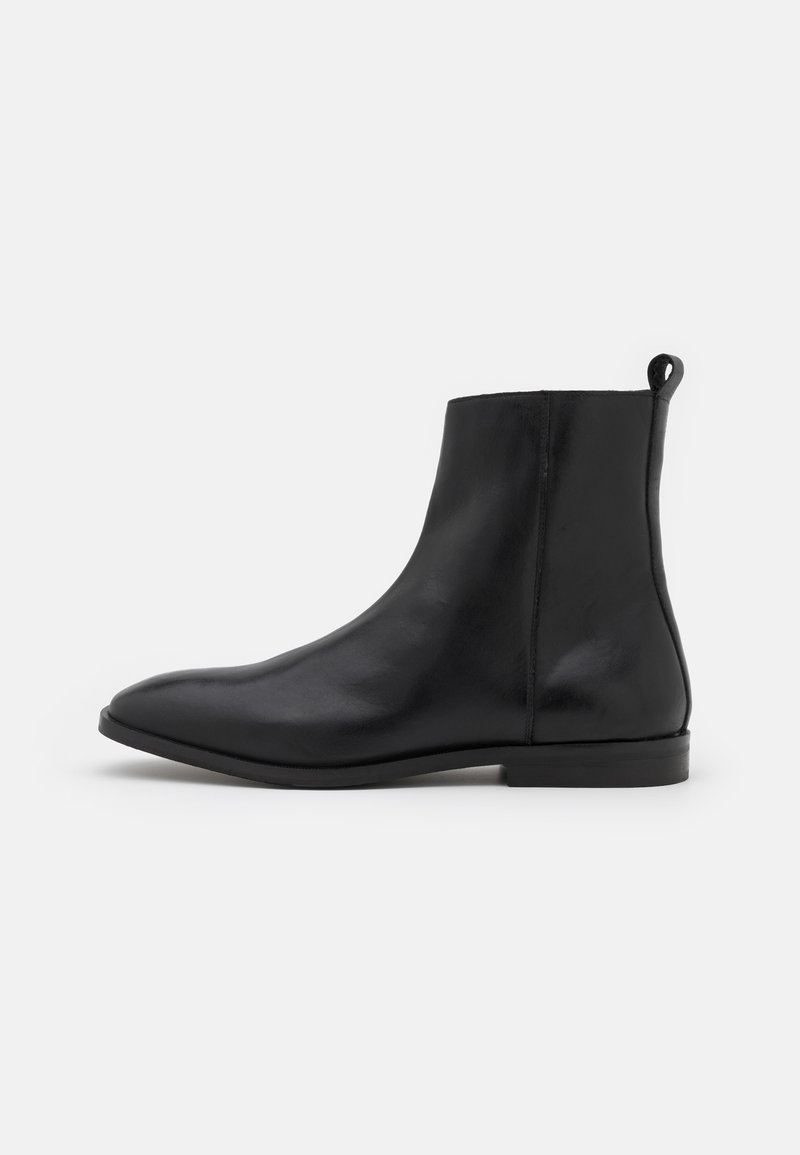 Bottine noire en cuir lisse avec une semelle plate et une languette à l'arrière, vue de profil sur un fond uni.
