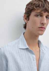 Jonge man met kort krullend haar, gekleed in een lichtblauw gestreept shirt, die iets opzij kijkt tegen een effen witte achtergrond.