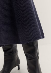 Gestreifte dunkelblaue Strickrock mit geribbelter Textur, kombiniert mit eleganten schwarzen Lederstiefeln bis zum Knie, die eine spitze Zehenform und einen dezenten Stilettoschuhabsatz haben.