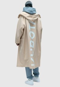 Beżowy wodoodporny płaszcz z białym napisem na plecach, w połączeniu z niebieską bluzą z kapturem, szarymi spodniami i białymi trampkami. Gładka tekstura, oversize'owy krój.