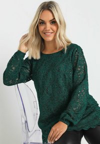 Grön spets långärmad topp med blommönster och paljetter, med rund hals och rynkade ärmkläppar, bärs över åtsittande svarta byxor.