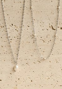 Dos collares de plata sobre un fondo claro texturizado; uno presenta un colgante de perla única, y el otro tiene pequeños acentos de perla a lo largo de la cadena.