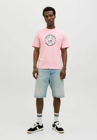 Rosa kurzärmliges T-Shirt mit einem schwarz-weißen Totenschädel-Graphic, kombiniert mit hellblauen Denim-Shorts und schwarzen Sneakers mit weißen Akzenten.