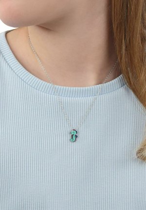 Collar de cadena plateada con colgante de caballito de mar turquesa y azul, llevado sobre un suéter acanalado azul claro por una persona de piel clara.