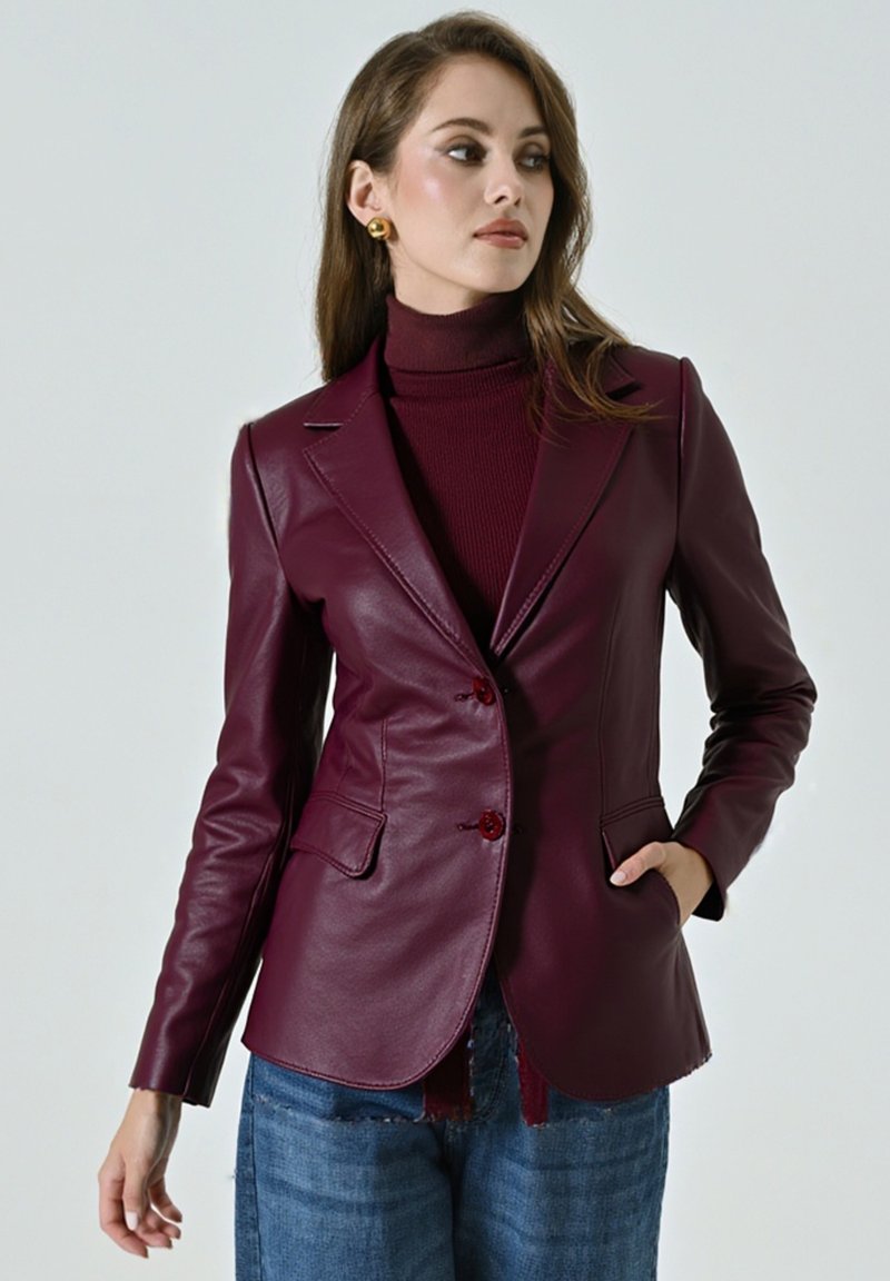 Donna che indossa un blazer in pelle bordeaux sopra un dolcevita abbinato, con jeans in denim blu, in posa con una mano in tasca e guardando di lato.