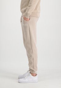 Beige bomulds sweatpants med elastisk talje og manchetter, med sidelommer og et hvidt "Alpha Industries" logo på låret.