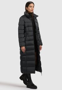 Sort puffet jakke med en lang silhuet, quiltet mønster, høj krave og sidelommer. Båret over et tætsiddende sort outfit og knæhøje støvler.