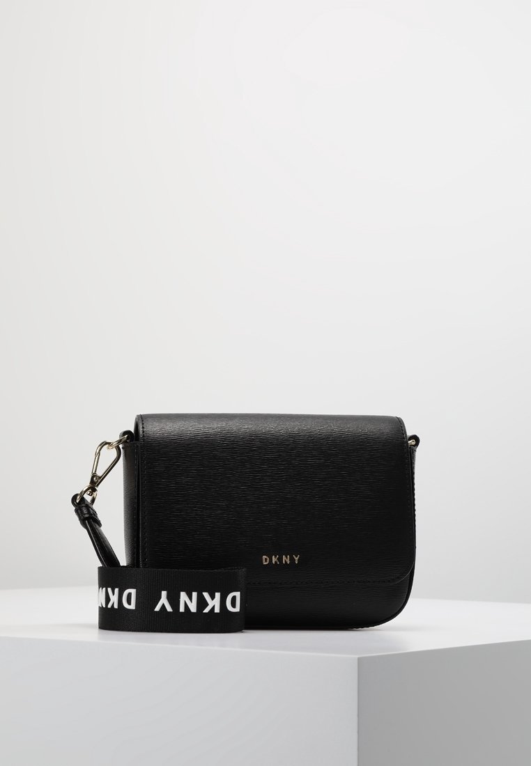 Sac bandoulière DKNY en cuir noir avec rabat magnétique et bandoulière en tissu de marque amovible reposant sur un piédestal blanc.