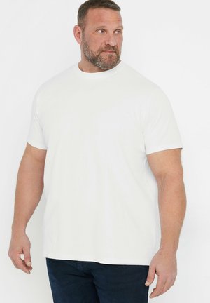 Erwachsener Mann mit kurzen Haaren und Bart, der ein einfarbiges weißes T-Shirt mit kurzen Ärmeln und dunkle Hosen trägt und nach links schaut.