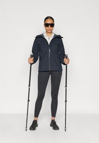 Tmavomodrá nepremokavá bunda so sivou kapucňou, doplnená o čierne legíny a tmavú obuv. Osoba drží turistické palice a má na sebe čierne slnečné okuliare.