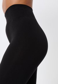 Leggings noirs taille haute en tissu extensible ; texture lisse sans motifs ni accessoires visibles. Gros plan sur le profil latéral.