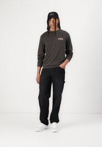 Dark gray sweatshirt with a round neckline and embroidered logo, paired with loose black jeans and white sneakers. Black cap with text.