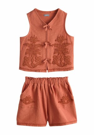 Meisje met lang zwart haar dat een mouwloze, roestkleurige top met borduursels en bijpassende korte broek draagt, staand en glimlachend met handen in de zakken.