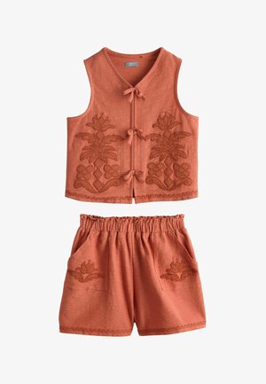 Fille aux longs cheveux noirs portant un haut sans manches couleur rouille brodé et un short assorti, debout et souriante avec les mains dans les poches.