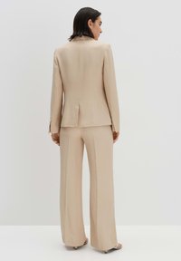 Completo in beige composto da un blazer aderente con uno spacchetto posteriore e pantaloni a gamba larga, realizzati in un tessuto morbido e liscio.