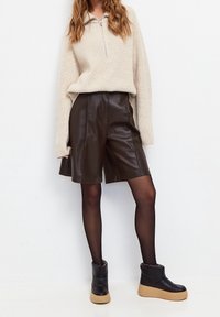 Mujer vestida con un suéter beige de cremallera, pantalones cortos de cuero marrón oscuro, medias negras y botines de plataforma negros con suelas beige.