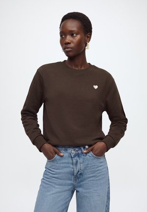 Femme aux cheveux courts portant un sweat-shirt marron foncé avec un petit cœur blanc sur la poitrine et un jean bleu clair, mains dans les poches.