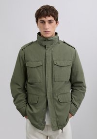 Young man wearing an olive green military-style jacket with four flap pockets and hands in side pockets, standing against a plain background.