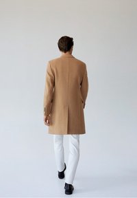 Abrigo de lana marrón claro con un diseño a medida, con un alto cuello y una abertura en la parte trasera, combinado con pantalones blancos y zapatos negros.