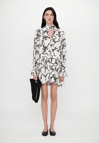 Rochie albă cu imprimeu abstract în formă de fundă neagră, decolteu în V, mâneci lungi, talie strânsă, fustă evazată; modelul ține o poșetă texturată neagră.