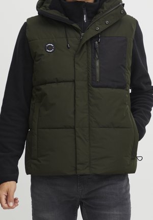 Mand iført en olivengrøn hættetrøje med sort brystlomme og sort langærmet trøje, hænder afslappet ned langs siderne.