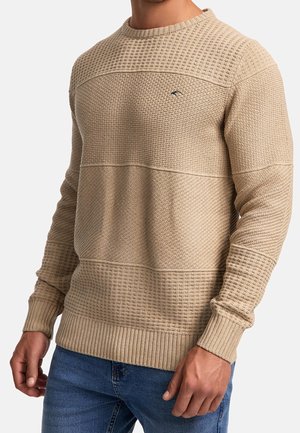 Mand iført en beige tekstureret striktrøje med ribbede manchetter og kant, kombineret med blå jeans, mod en ensfarvet baggrund.