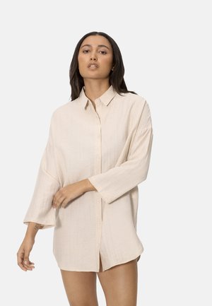 Mujer con cabello largo y oscuro que lleva una camisa suelta de color beige claro con botones y mangas tres cuartos, contra un fondo liso.