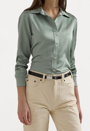 Femme portant une chemise boutonnée manches longues vert clair et un pantalon beige avec broderies florales, accessoirisée d'une ceinture noire avec une boucle brillante.